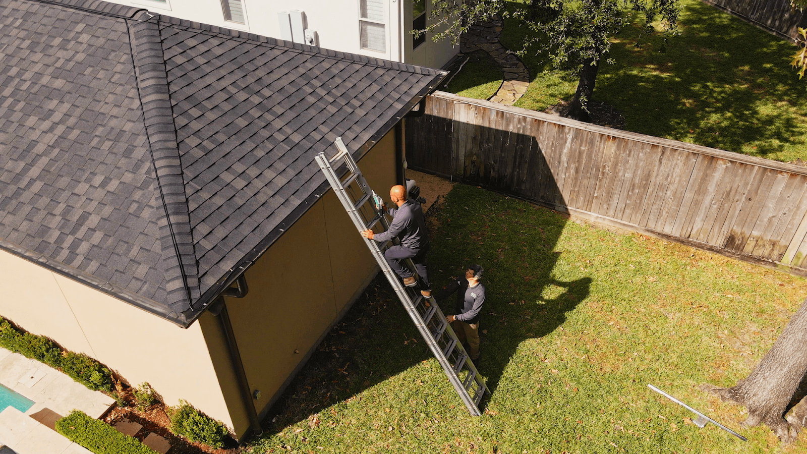 Two technicians inspect and seal roofline entry points from an extension ladder on a suburban home.
