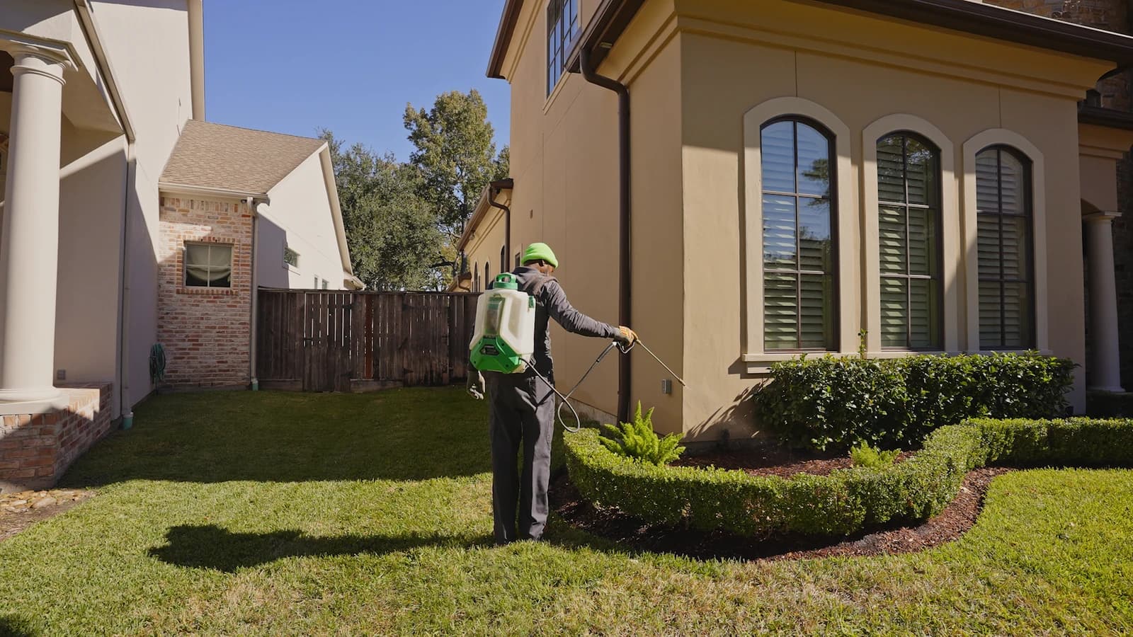 Licensed technician in a bright green shirt sprays liquid pest treatment along the brick foundation of a two-story home.