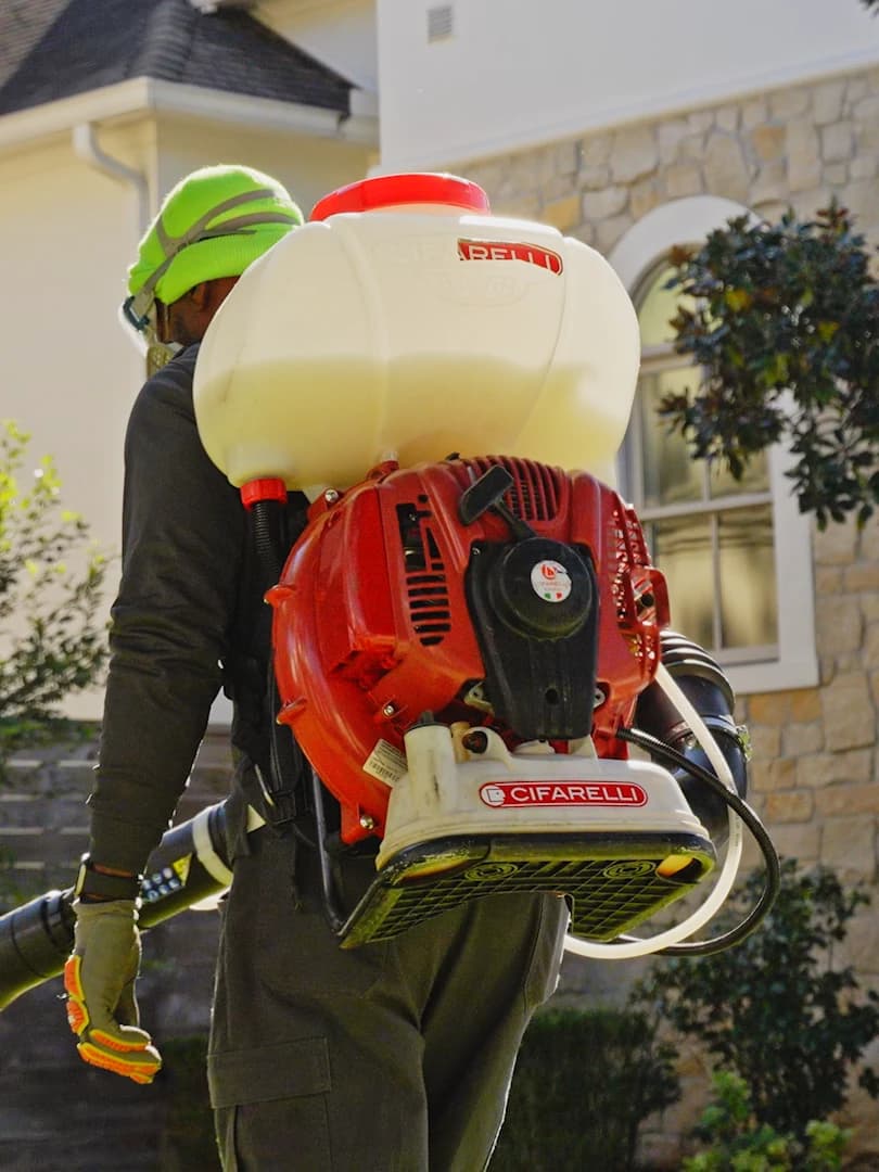 Pest control technician in protective gear carries a backpack mist blower toward the landscaping of a stone-and-stucco house.